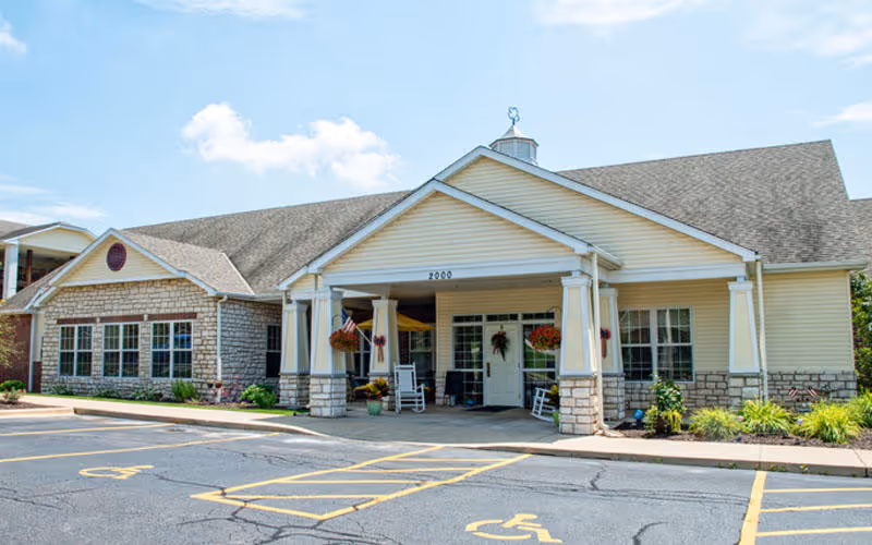 Front exterior view of a single-story senior living facility building with a covered entrance supported by columns, hanging flower baskets, rocking chairs on the porch, and a parking lot with handicap spaces in front under a partly cloudy sky.