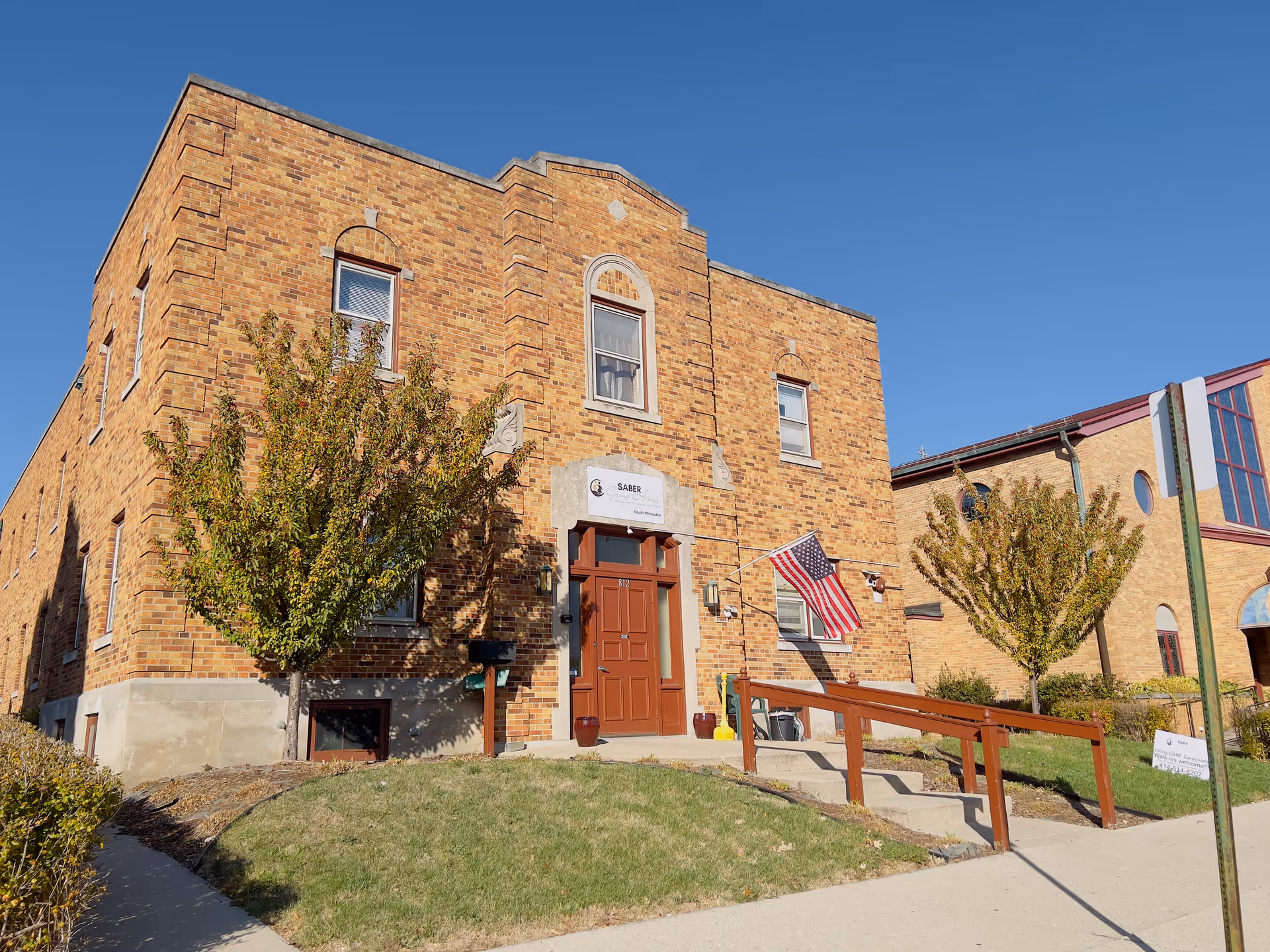 Brick two-story building front with an entrance ramp, American flag, and small trees under a clear blue sky.