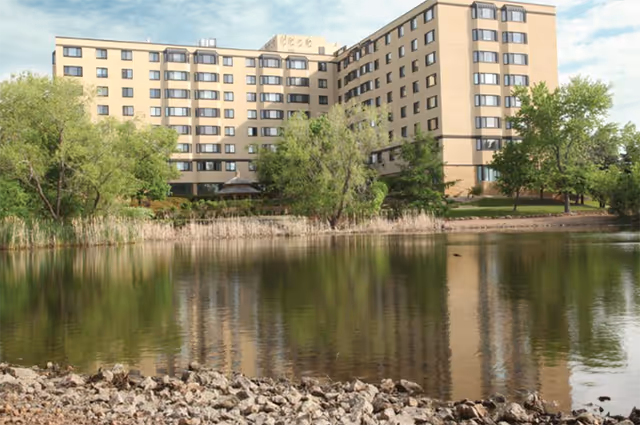 Multi-story apartment building reflected in a pond with trees and a rocky shoreline in the foreground.