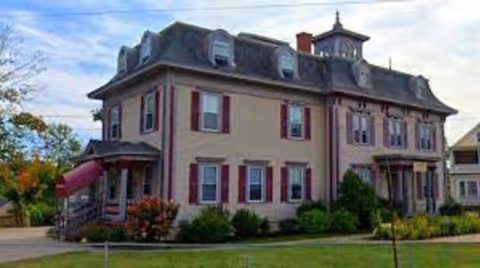 A large, two-story historic building with beige walls and red shutters, surrounded by green shrubs and a lawn under a partly cloudy sky.