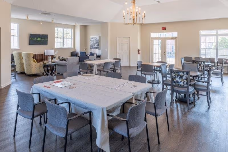 A spacious senior living facility common area with multiple tables and chairs arranged for group activities or dining. The room has large windows allowing natural light, a chandelier, and a seating area with armchairs and a side table. A sign on the wall reads 'Amara Place'.