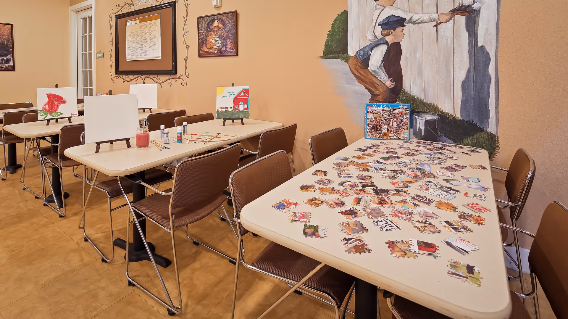 A room with several tables and chairs arranged for activities. One table has a partially completed jigsaw puzzle with scattered pieces, and another table has blank canvases on easels along with paintbrushes and paint bottles. The walls are decorated with paintings and a mural of two children painting a fence.
