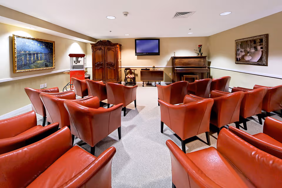 A cozy room with multiple red leather armchairs arranged in rows facing a wall-mounted flat screen TV. The room also features a wooden piano, a wooden cabinet, a popcorn machine, and framed artwork on the walls. The lighting is warm and the carpet is light-colored.