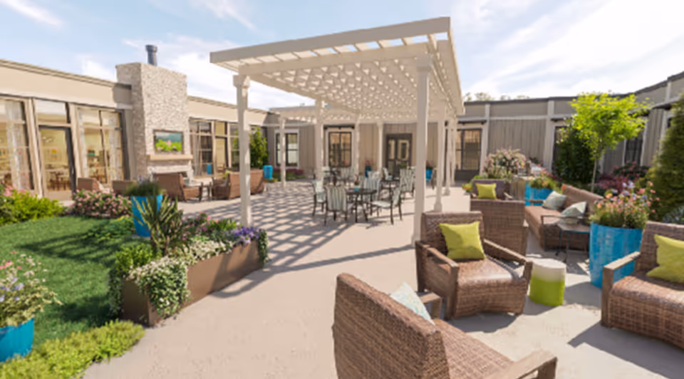 Outdoor courtyard area at Calyx Living of Fuquay-Varina featuring a shaded pergola with tables and chairs underneath, surrounded by wicker seating with colorful cushions, potted plants, and greenery, adjacent to building windows and doors.