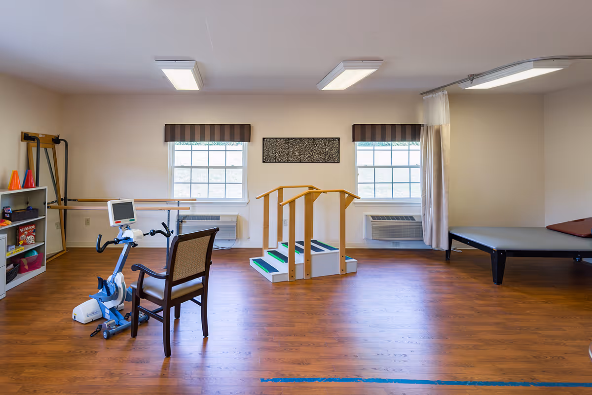 A spacious room with wooden flooring featuring physical therapy equipment including a stationary exercise bike with a chair, a set of stairs with handrails for rehabilitation, a therapy table, and shelves with various therapy tools and games. Two windows with striped valances allow natural light into the room.