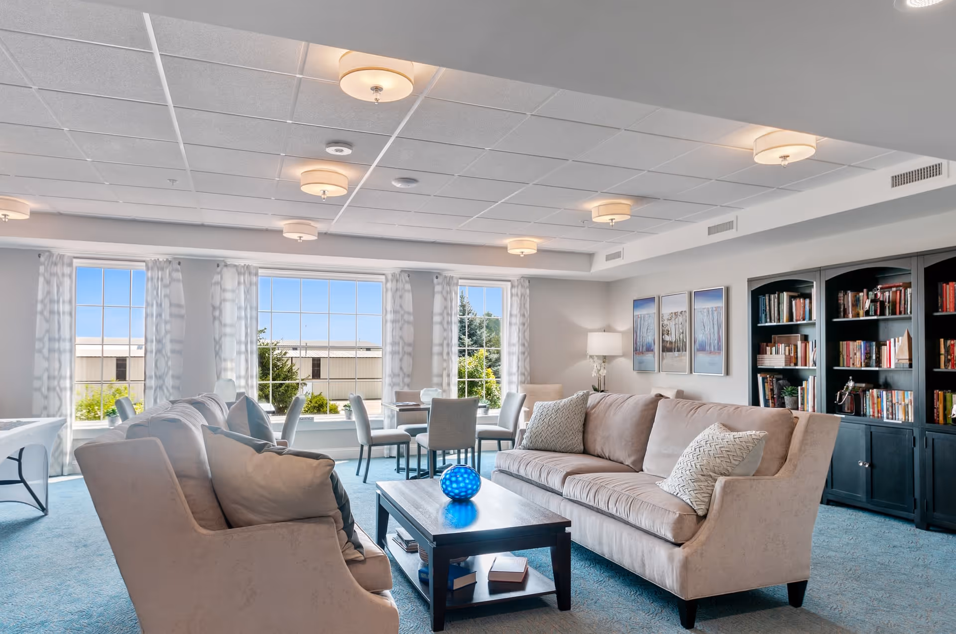 A bright and spacious living room with large windows letting in natural light. The room features two beige sofas with decorative pillows, a dark wooden coffee table with books and a blue decorative object, a set of dining tables and chairs near the windows, a floor lamp, framed artwork on the wall, and a large dark wooden bookshelf filled with books.