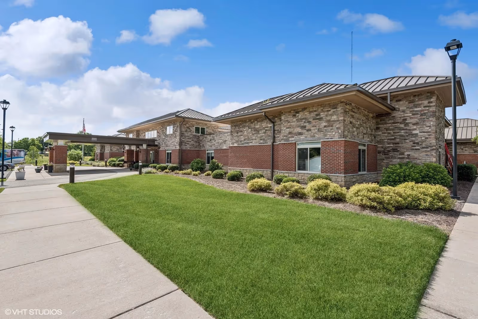 Exterior view of The Pearl at Watkins Centre - Memory Care Community building with a well-maintained lawn, shrubs, and a covered entrance under a partly cloudy sky.