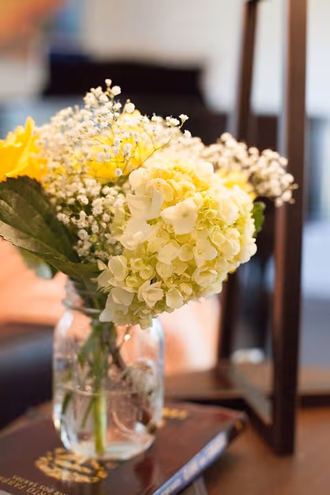 A close-up of a glass jar filled with water holding a bouquet of flowers including white hydrangeas, yellow roses, and baby's breath, placed on top of a book on a wooden table with a blurred background.