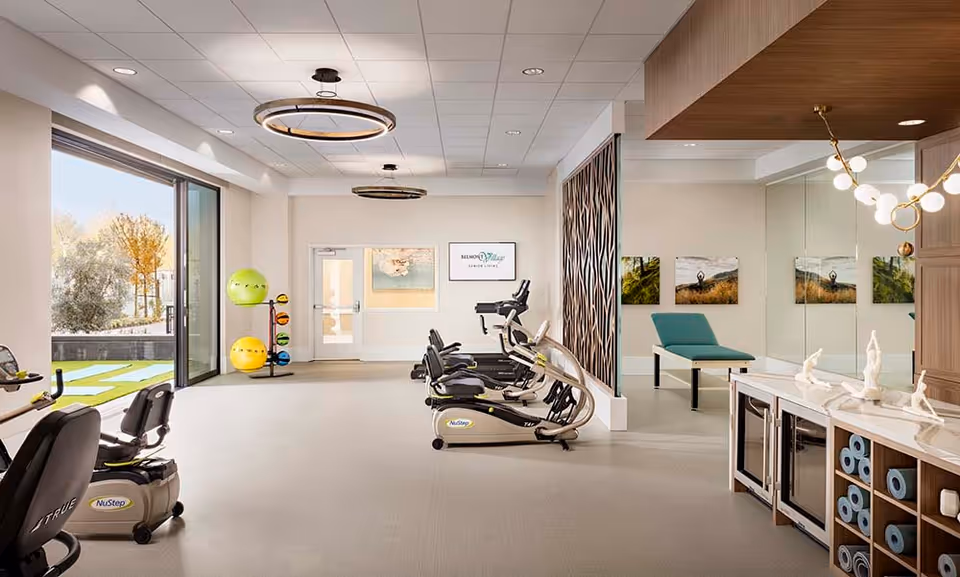 Bright and spacious fitness room at Belmont Village San Ramon with exercise bikes, elliptical machines, colorful exercise balls, and a large window showing an outdoor area. The room features modern lighting, a decorative partition, and a side area with a teal exercise bench and wall art.