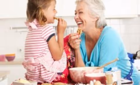 An elderly woman and a young girl smiling and enjoying cookies together in a bright kitchen setting with baking ingredients on the counter.