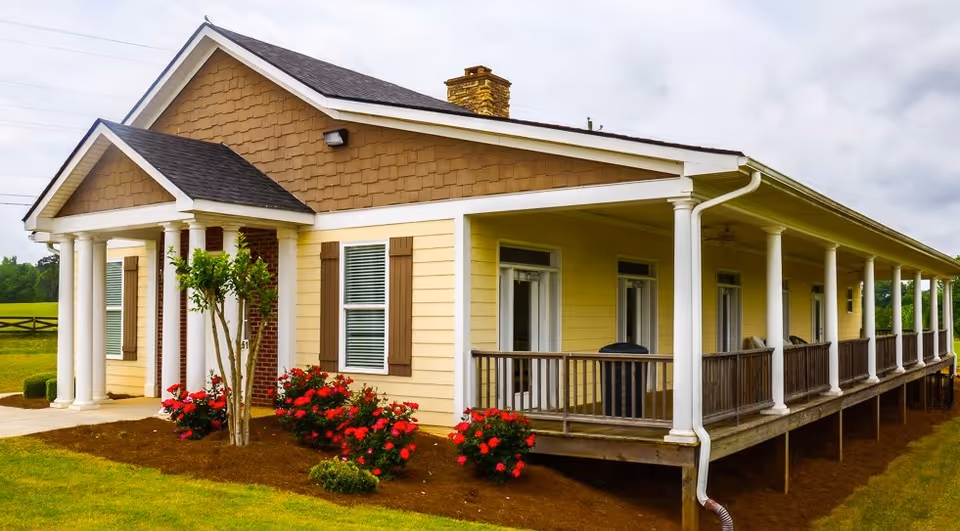 Exterior view of a single-story yellow building with a large wrap-around porch supported by white columns, surrounded by green grass and red flowering bushes under a cloudy sky.