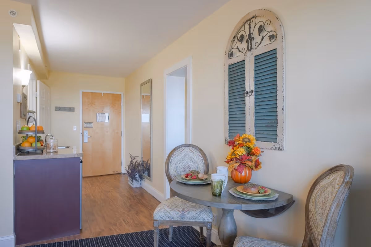 A small dining area in a senior living facility with a round table set for two, featuring plates with bagels and strawberries, a green drink, and a pumpkin vase with autumn flowers. The room has light-colored walls, wooden flooring, a decorative wall piece with blue shutters, and two upholstered chairs. In the background, there is a kitchenette with a fruit stand and a wooden entrance door.