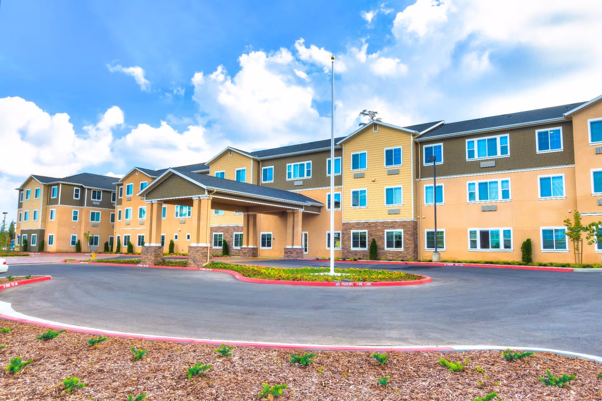 Exterior view of a large, multi-story senior living facility building with a covered entrance, surrounded by a circular driveway and landscaped areas under a partly cloudy blue sky.