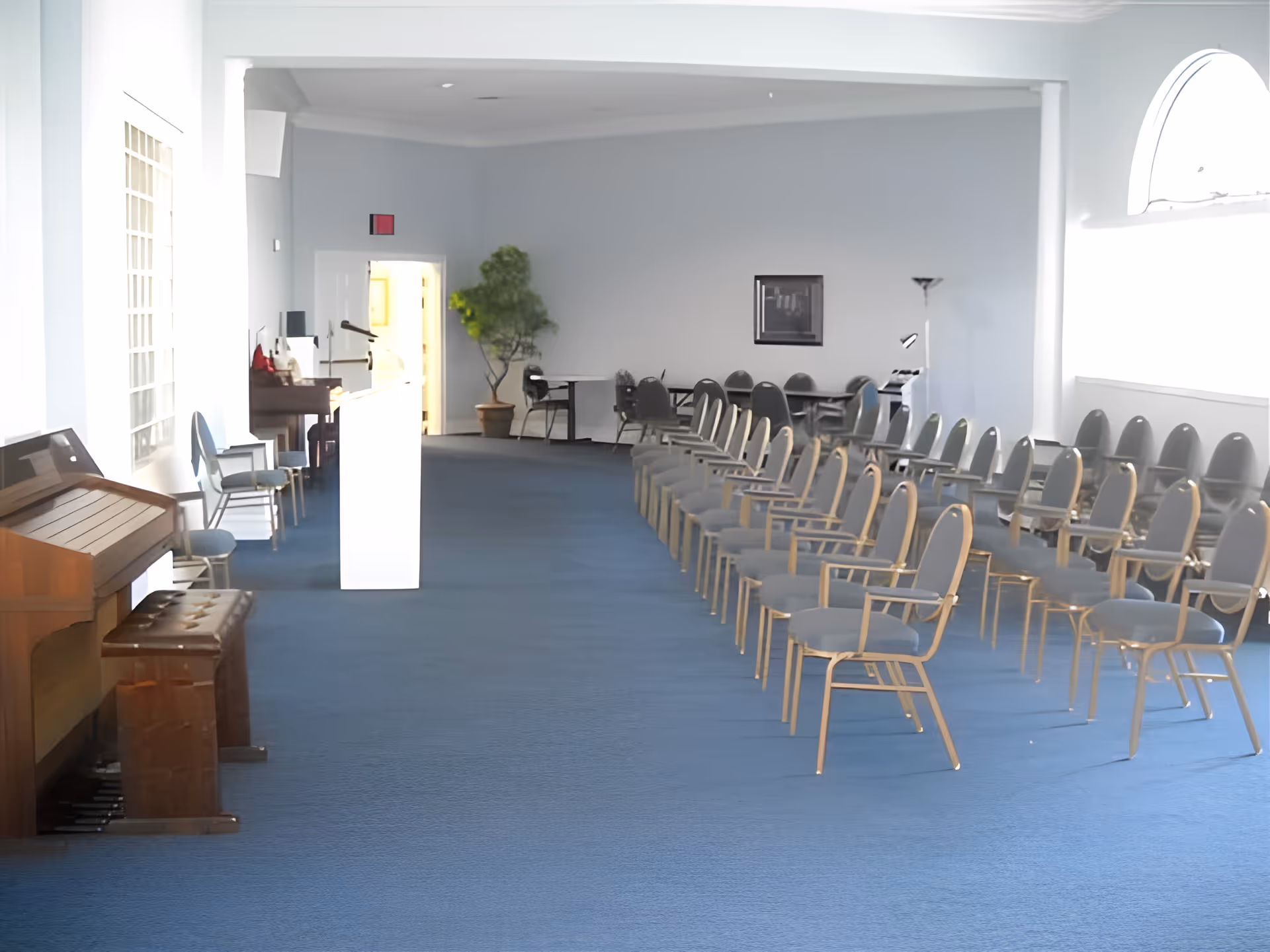Spacious meeting room with rows of chairs facing a podium, a piano at left, and blue carpet.