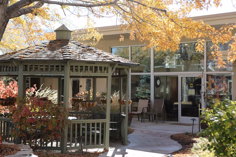 A green wooden gazebo with chairs and hanging planters in a landscaped courtyard outside a building entrance framed by autumn foliage.