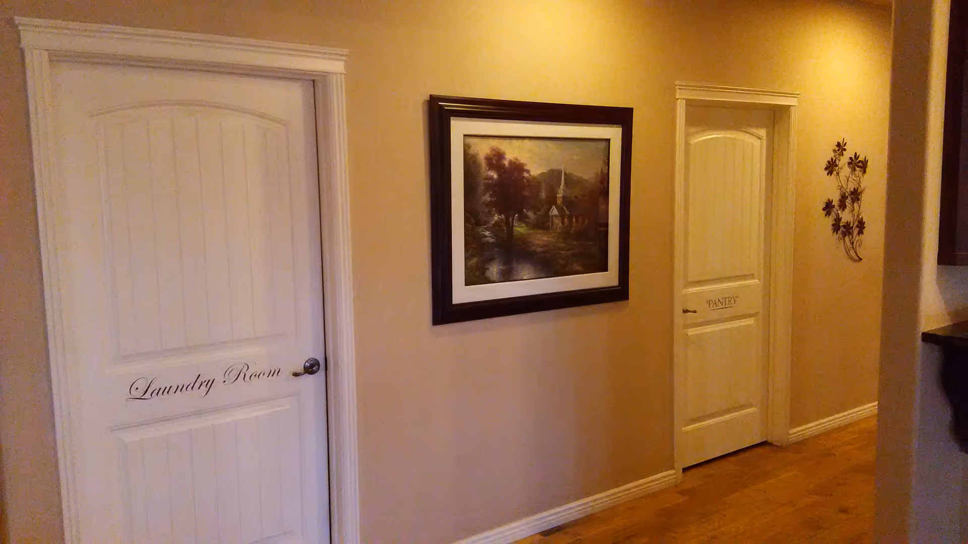 Interior hallway with two white doors labeled 'Laundry Room' and 'Pantry'. Between the doors is a framed painting of a scenic landscape with a church and trees. The walls are painted beige, and there is a wooden floor. A decorative metal wall art piece with flowers is mounted on the wall near the pantry door.