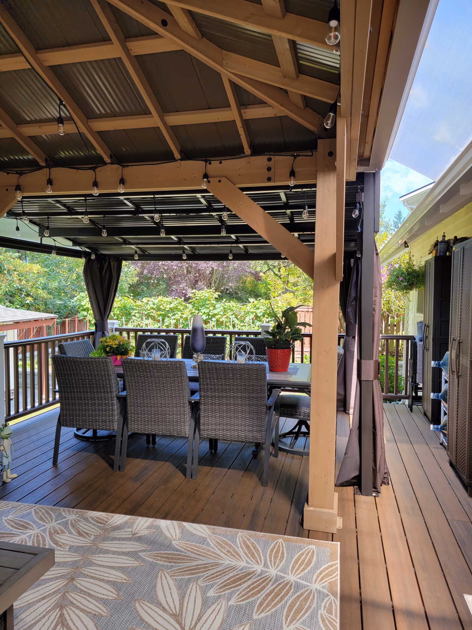 Covered outdoor dining patio with a wooden pergola, string lights, a table with wicker chairs, potted plants, and surrounding greenery.