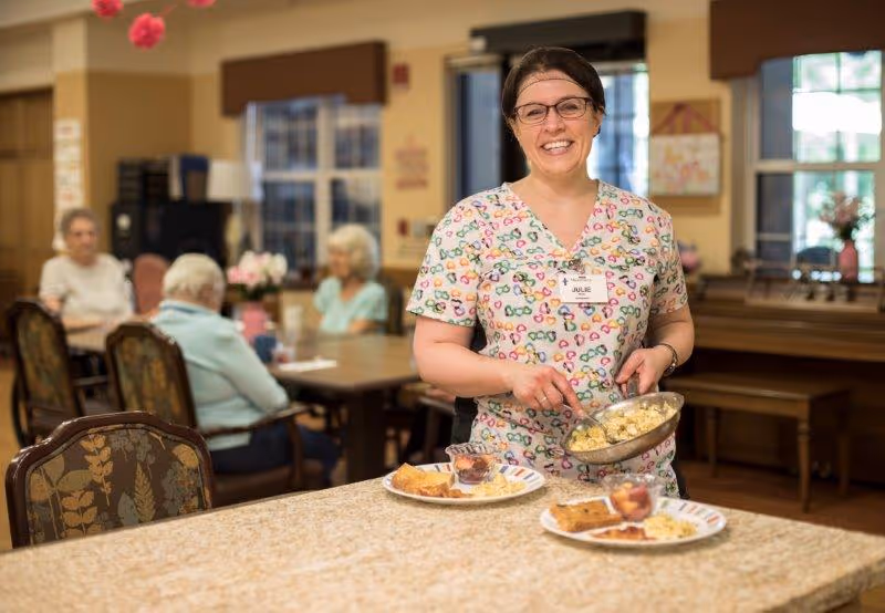 A smiling staff member serves plated breakfast food in a senior living dining room with residents seated at tables in the background.