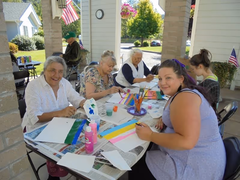 A group of elderly women and a younger woman sitting around a table outdoors under a covered patio, engaged in painting and arts and crafts activities. The table is covered with newspapers and various painting supplies including brushes, paint bottles, and paper. American flags and greenery are visible in the background.