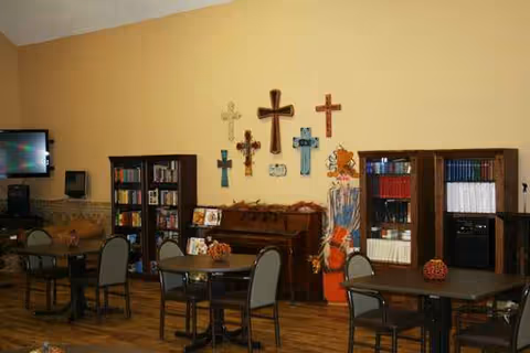 Interior communal room at Hemphill Care Center with tables, chairs, bookshelves, piano, and wall crosses.