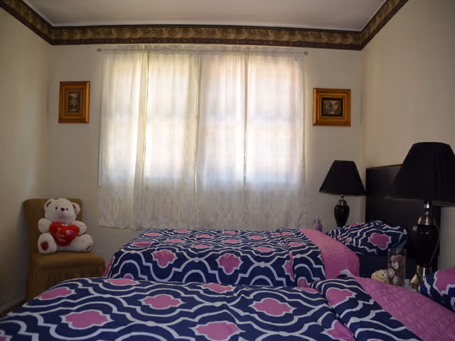 Two twin beds with blue and pink patterned bedding in a softly lit bedroom with a window, bedside lamps, and a teddy bear on a chair.