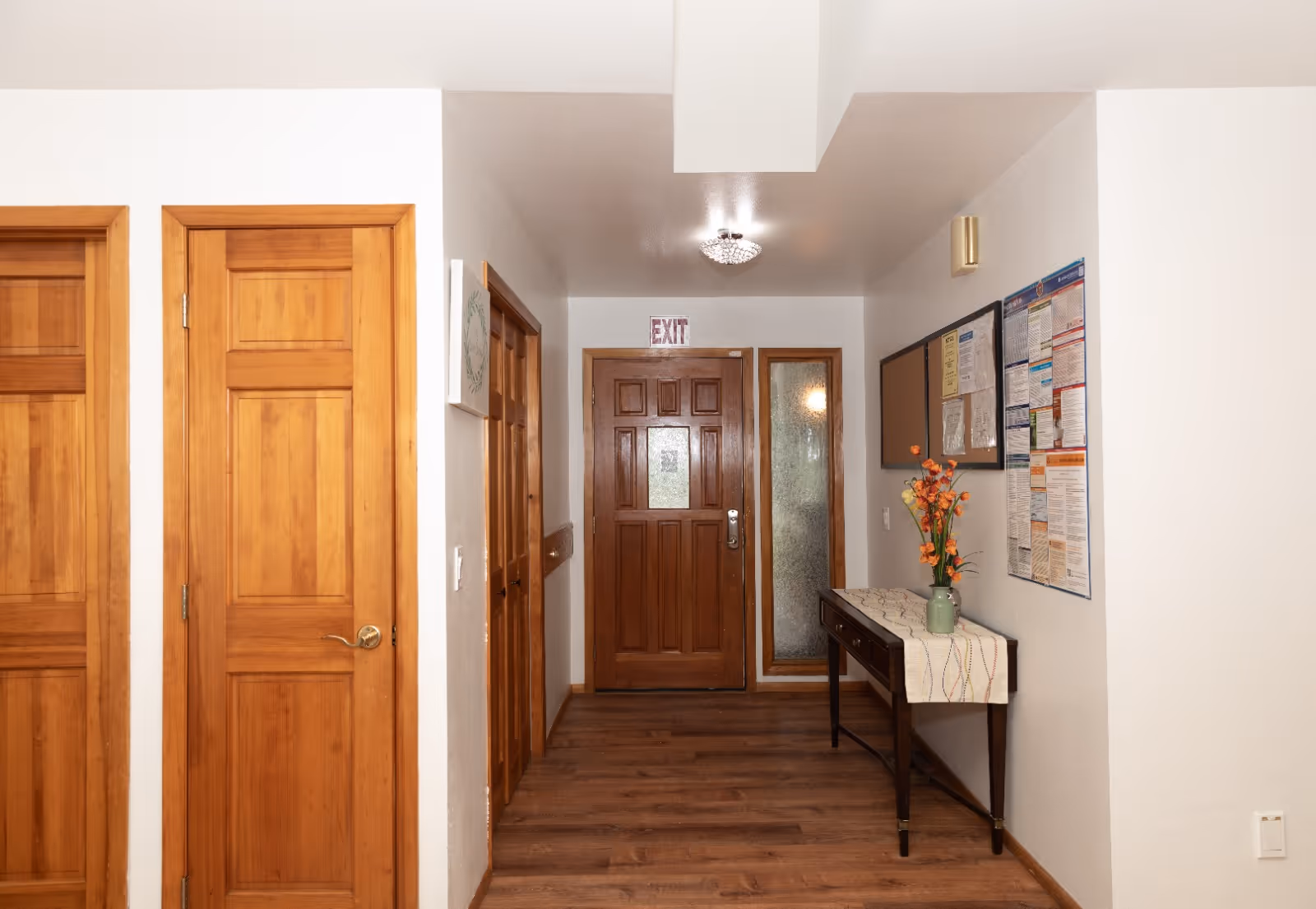 Interior hallway of Vistra Care Adult Family Home with wooden doors on the left, a wooden exit door straight ahead, and a narrow frosted window beside it. On the right wall, there is a small table with a vase of orange flowers and a table runner, along with a bulletin board displaying various notices.
