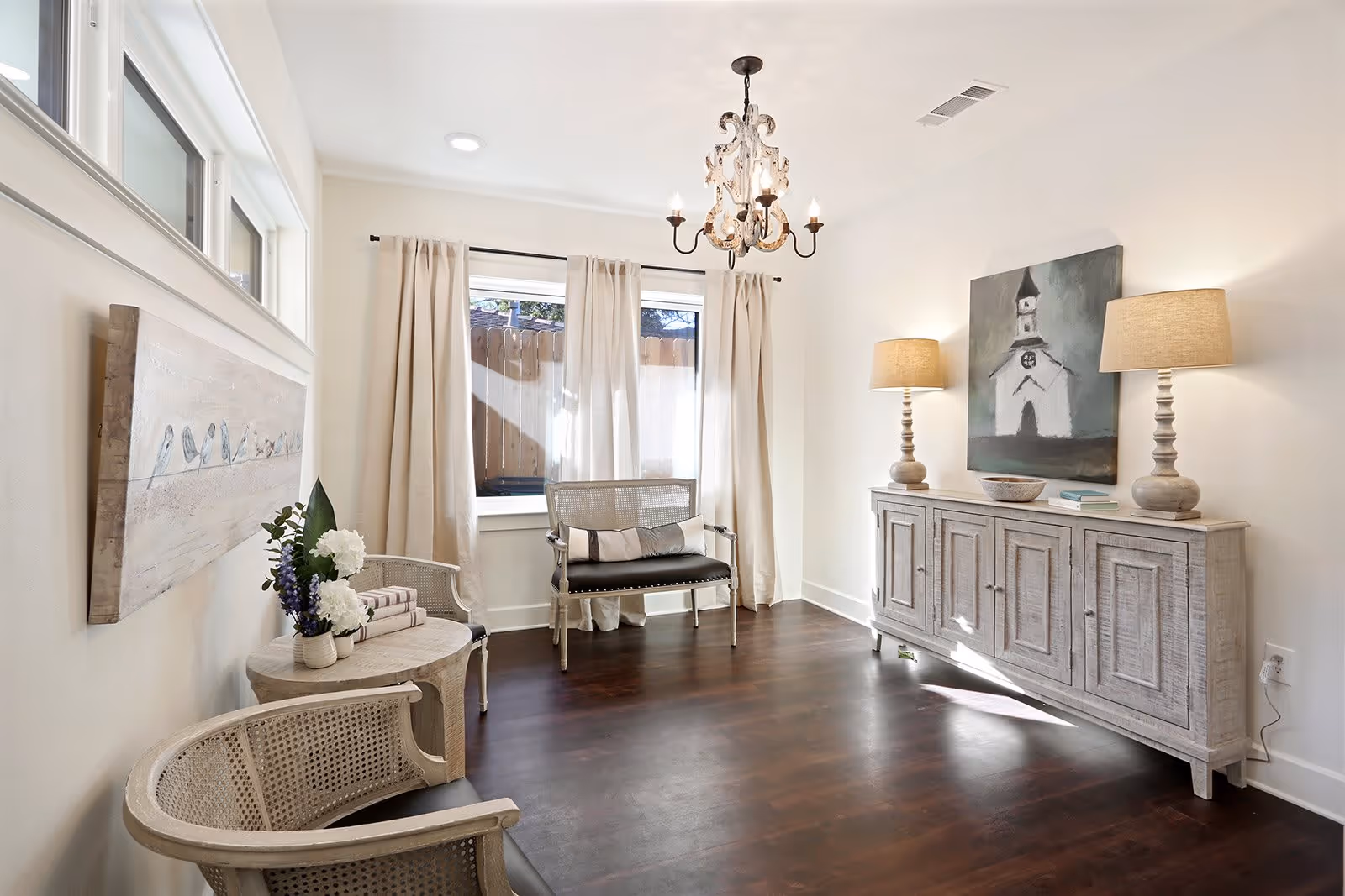 Bright sitting room with dark wood floors, light-colored chairs and sideboard topped with lamps, a chandelier, and a curtained window.