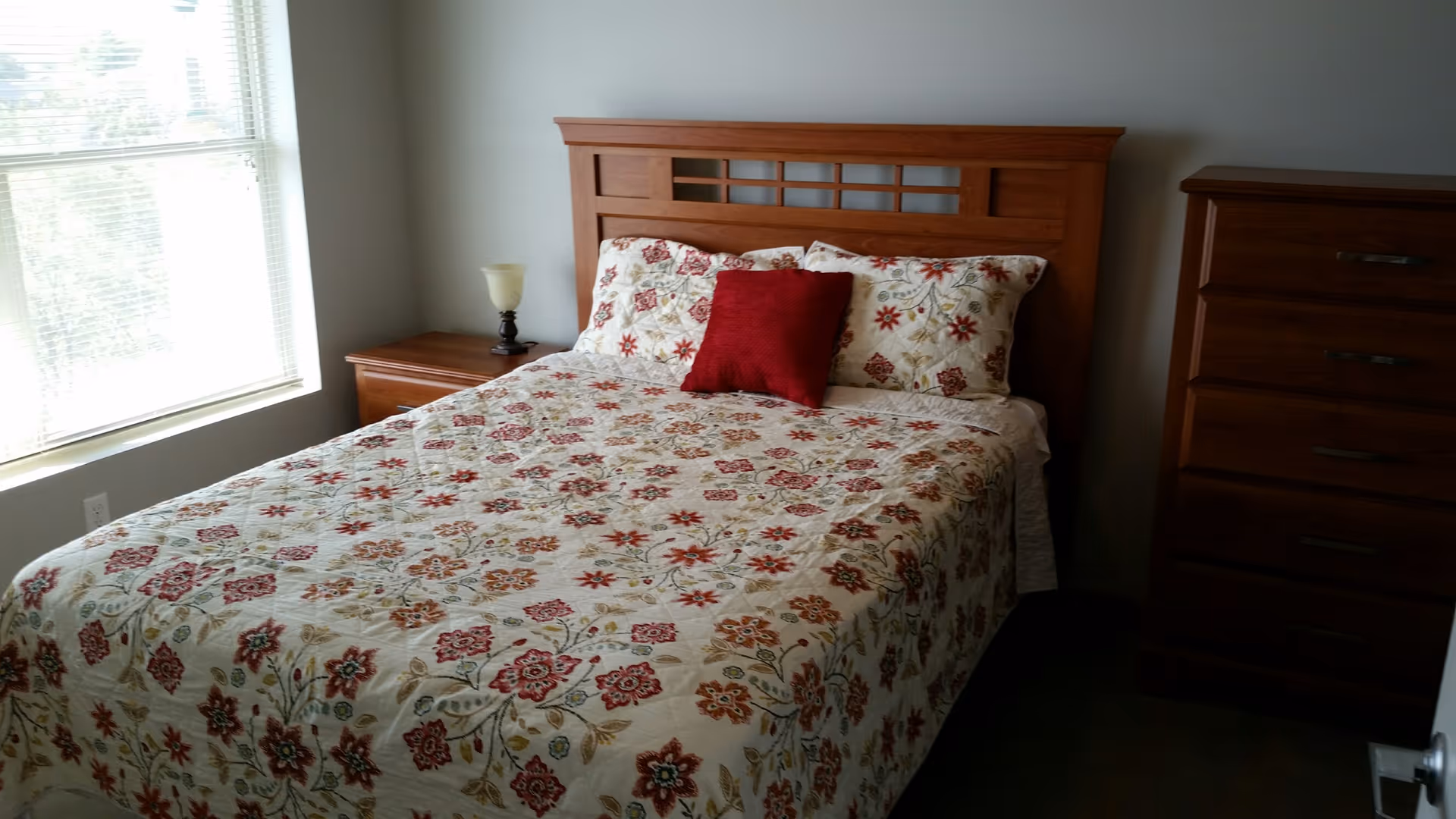 A bedroom with a wooden bed frame and a floral patterned bedspread featuring red, orange, and green flowers. There are two pillows with matching floral pillowcases and a single red accent pillow. To the left of the bed is a wooden nightstand with a small lamp, and to the right is a tall wooden dresser. A window with blinds is on the left wall, allowing natural light into the room.