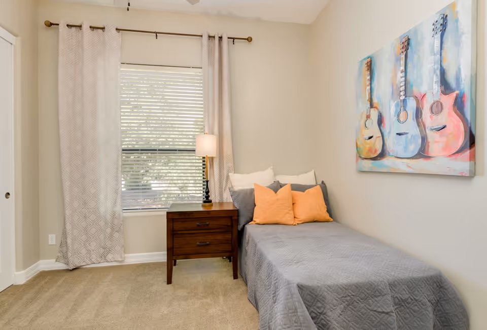 A small bedroom with a single bed covered in a gray quilt and decorated with orange and gray pillows. Next to the bed is a wooden nightstand with two drawers and a lamp on top. A window with closed blinds and beige patterned curtains is behind the nightstand. On the wall above the bed is a colorful painting of three guitars.