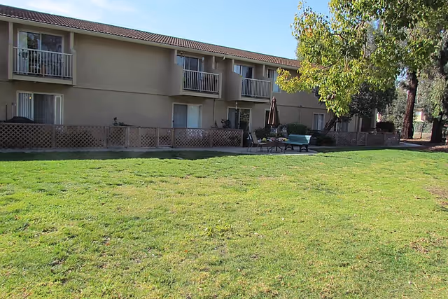 Exterior view of a two-story retirement community building with balconies and sliding glass doors. In front of the building is a large grassy lawn with a patio area featuring a table, chairs, an umbrella, and a bench. Trees and shrubs surround the area under a clear sky.