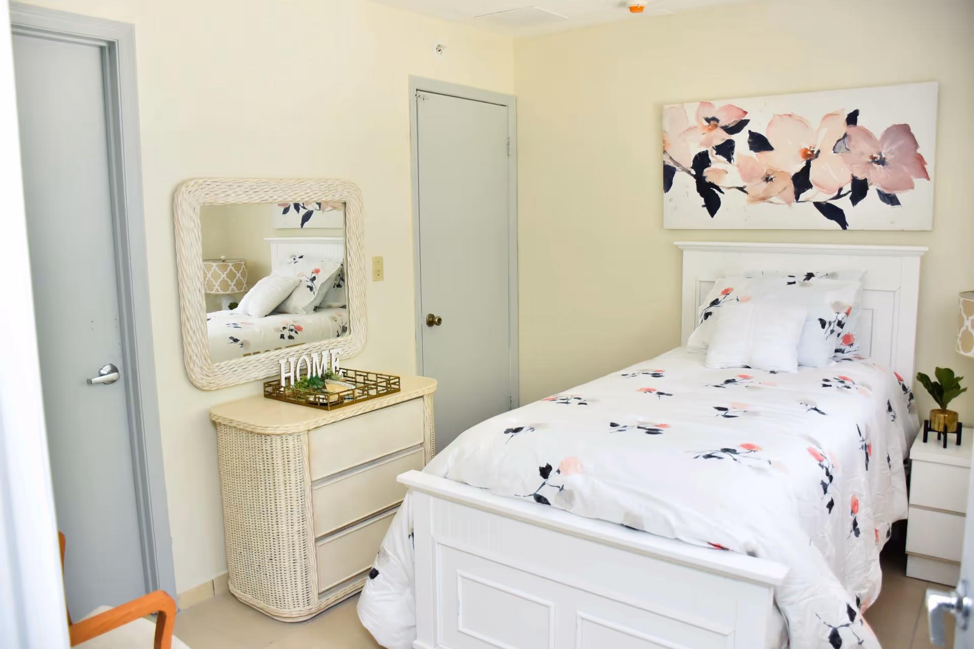 Bright single bedroom with a white wooden bed dressed in floral bedding, a wicker dresser with mirror, a nightstand, and floral wall art.