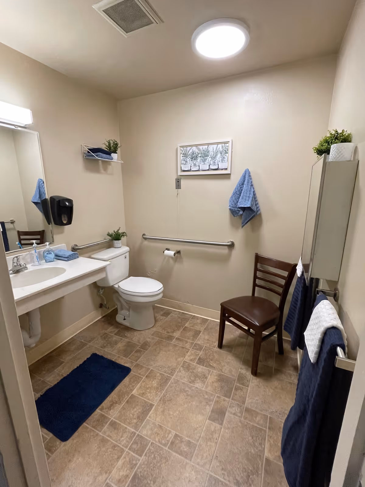 A clean and accessible bathroom with beige walls and tiled floor. The room features a white toilet with a grab bar on the wall beside it, a white sink with a countertop, a large mirror, and a black soap dispenser. There is a wooden chair with a brown seat, blue towels hanging on hooks and a towel rack, a small shelf with a plant and folded towels, and a framed picture on the wall. A blue bath mat is placed on the floor.