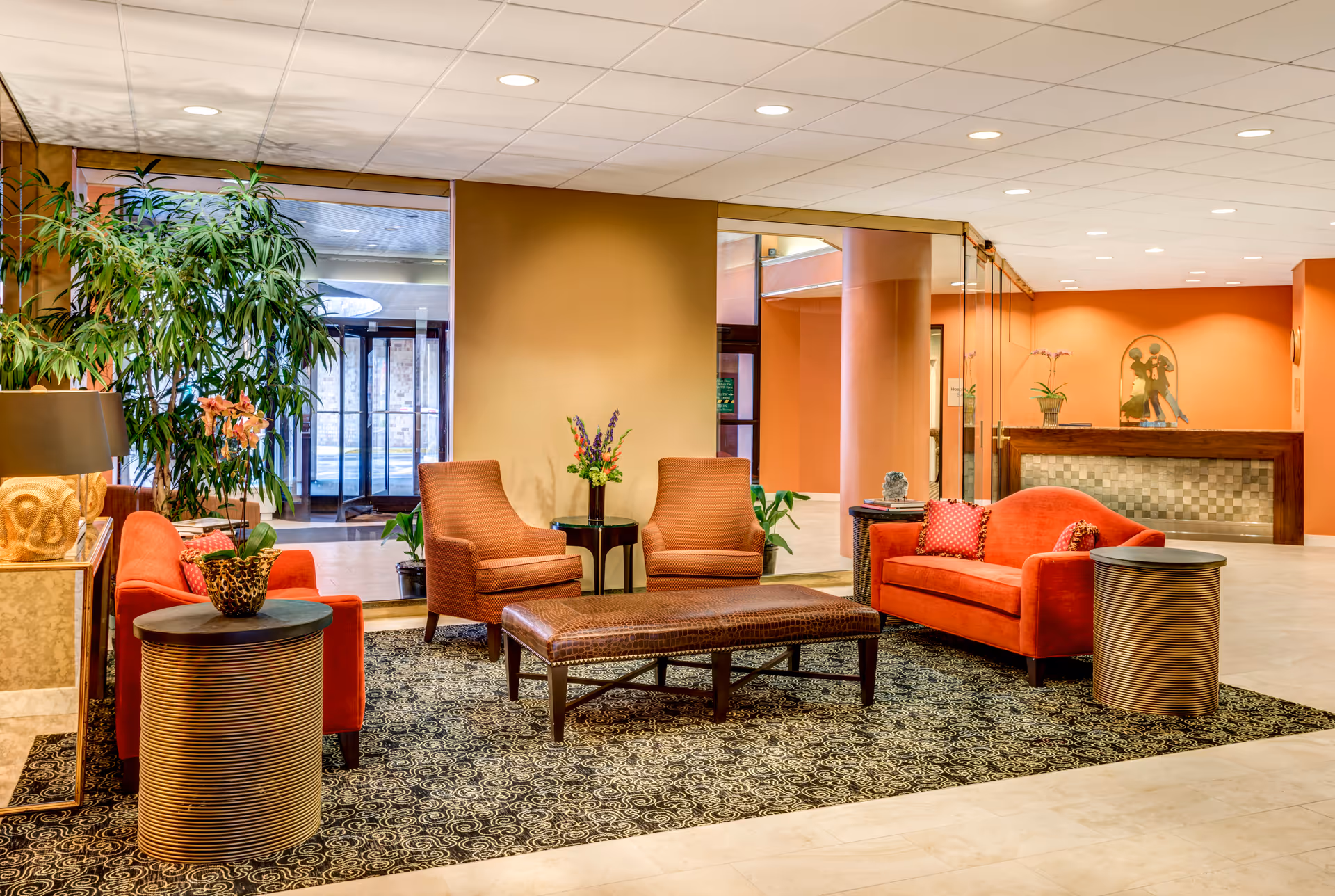 A cozy seating area in a senior living facility lobby with two orange sofas, two brown armchairs, a large rectangular ottoman, and two round side tables. The space features a patterned rug, potted plants, warm orange walls, and a reception desk in the background.