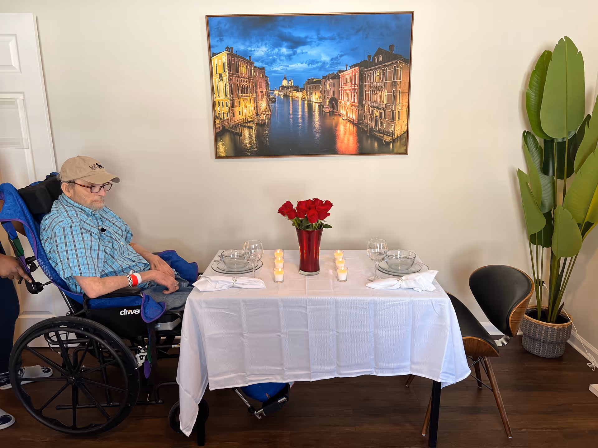 An elderly man in a wheelchair sits next to a dining table set for two with a white tablecloth, glass plates, wine glasses, napkins, and a red vase with red roses in the center. There are small lit candles on the table. A large framed picture of a canal scene at dusk hangs on the wall behind the table. A tall green plant in a woven basket is placed to the right of the table.