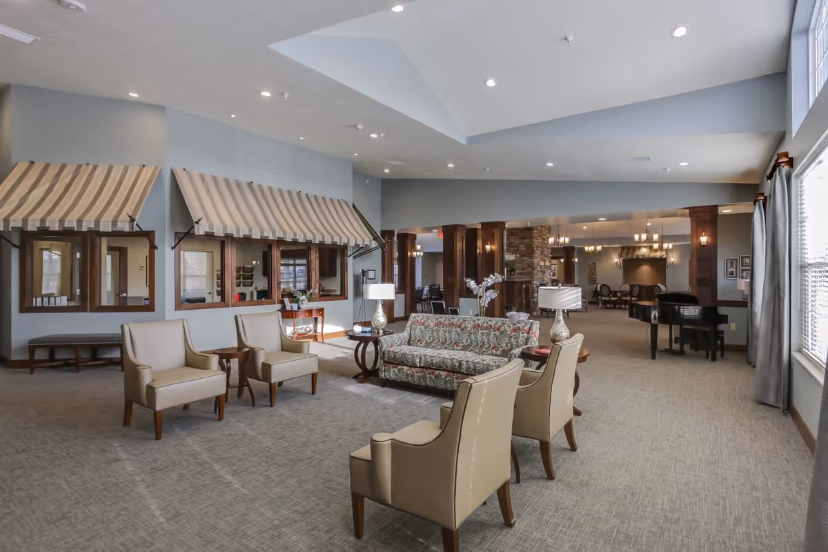A spacious and well-lit living room area in an assisted living facility featuring a patterned sofa, four beige armchairs arranged around two small tables with lamps, large windows with curtains on the right, and a piano in the background near a stone fireplace and dining area.