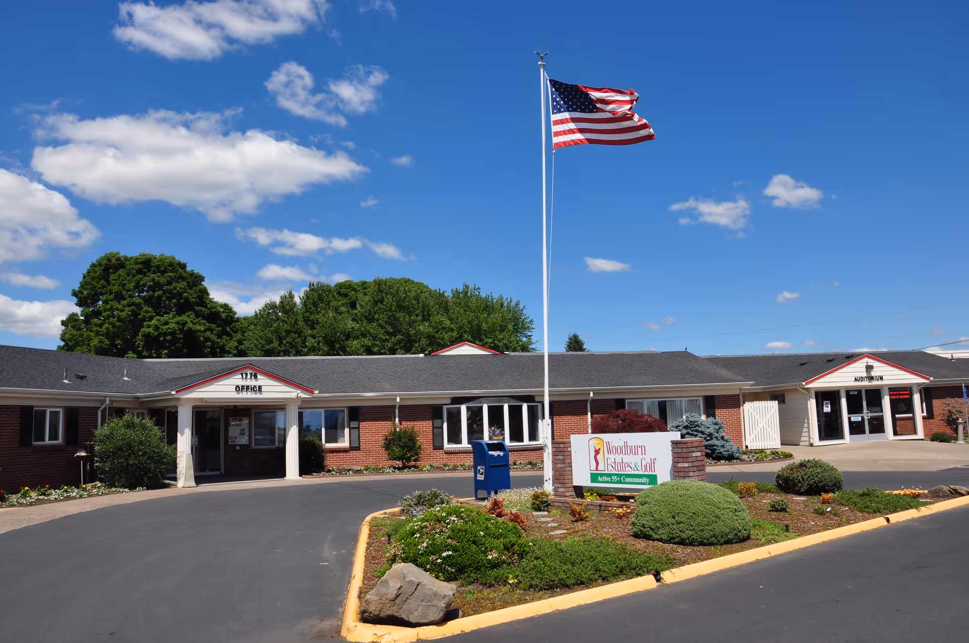 Single-story brick senior living facility with an American flag on a tall pole and a landscaped circular driveway with a sign.