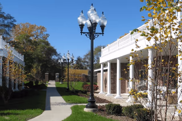 Outdoor walkway between two buildings with white columns and decorative street lamps under a clear blue sky, surrounded by green grass and trees with some autumn leaves.