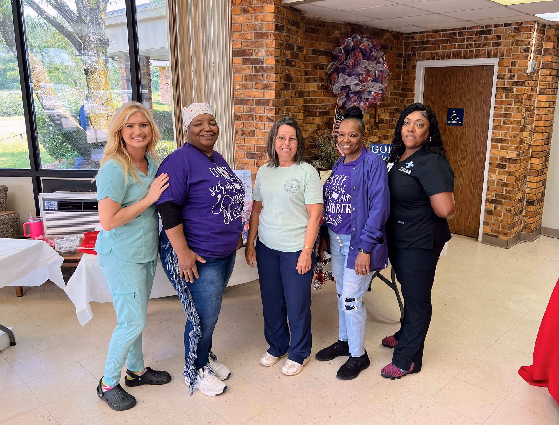 Five women standing together inside a facility with brick walls and large windows. They are smiling and posing for the photo. One woman is wearing light blue scrubs, two women are wearing purple shirts with text, one woman is wearing a light green shirt, and one woman is in black scrubs. There is a wreath decoration on the wall and a door with a handicap restroom sign behind them.
