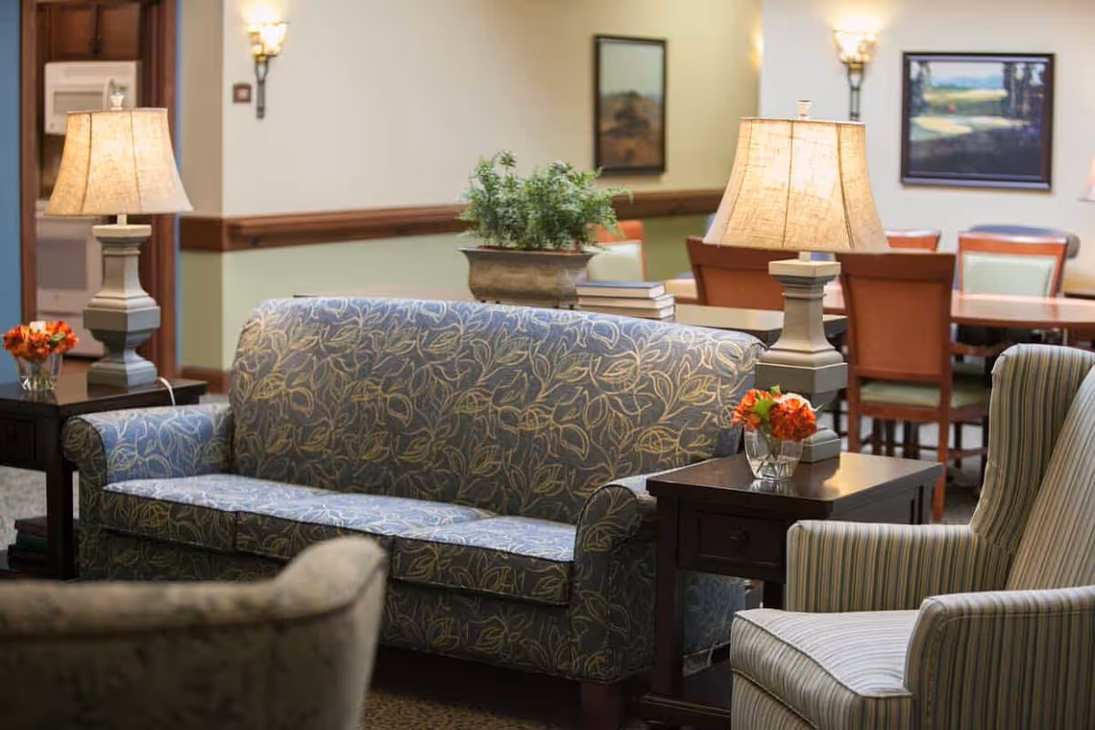 A cozy senior living common area with a patterned blue sofa, striped armchair, two wooden side tables with lamps, and small flower arrangements. In the background, there is a dining area with tables and chairs, wall sconces, and framed artwork on the walls.