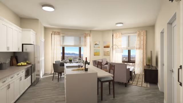 A bright and modern senior living facility interior featuring a kitchen area with white cabinets, a refrigerator, and a microwave on the left. In the center, there is a kitchen island with two stools, a bottle of wine, and glasses. Beyond the island, there is a dining table with chairs near large windows with beige curtains, offering a scenic view outside. To the right, there is a cozy living area with two armchairs, a floor lamp, a TV on a stand, and decorative wall art. The flooring is wood-style, and the walls are painted a light color.