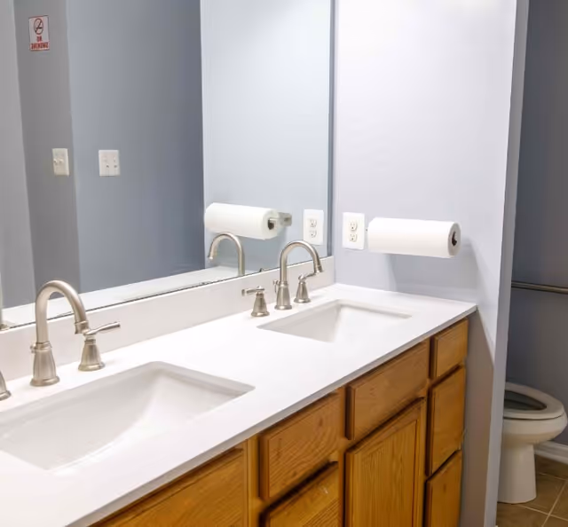 A bathroom with a double sink vanity featuring two silver faucets and wooden cabinets below. A large mirror spans the length of the vanity, reflecting a wall-mounted paper towel holder with a roll of paper towels. In the background, a toilet is partially visible. The walls are painted light gray.