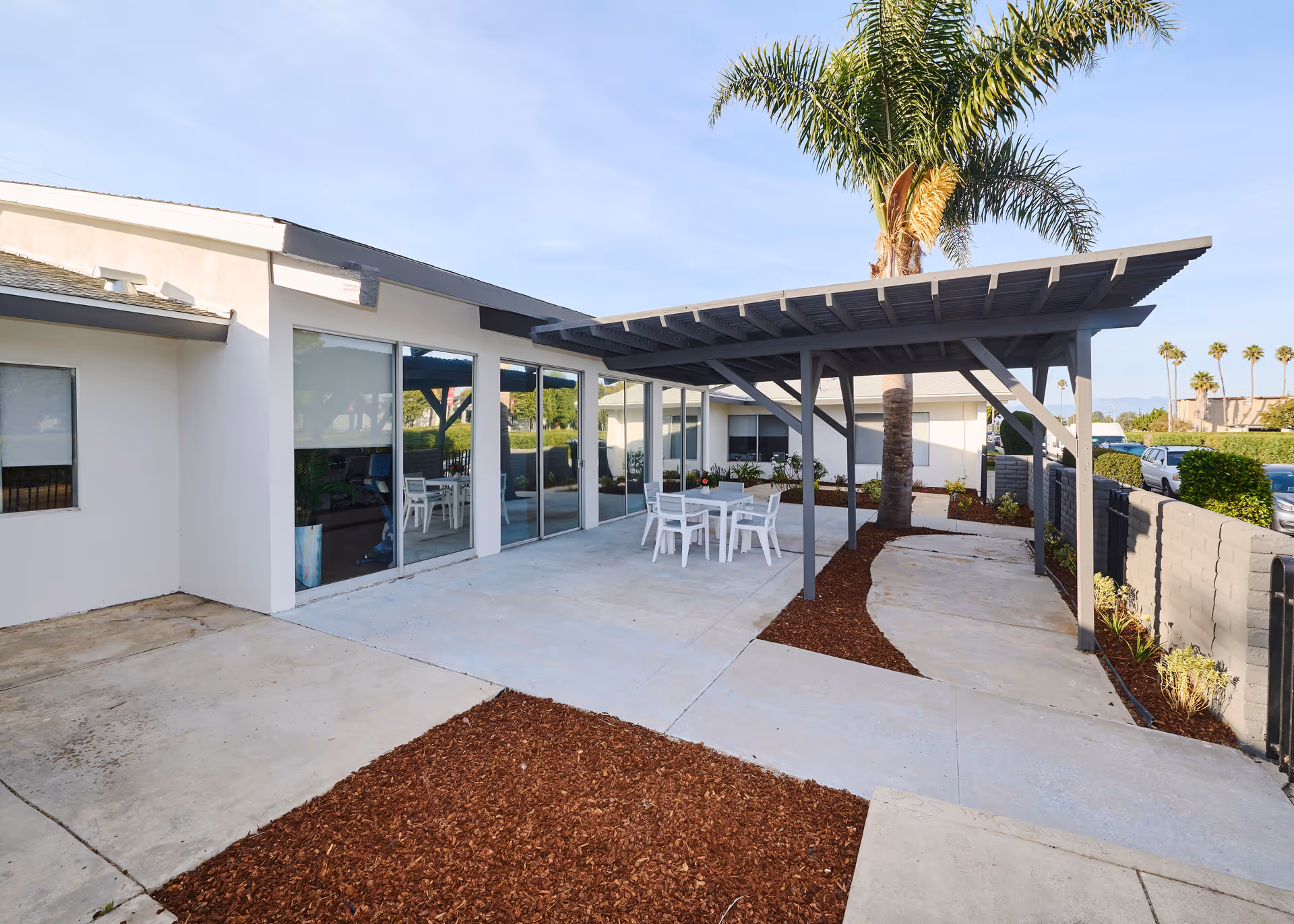 Outdoor patio area with a pergola, palm tree, and white table-and-chair set adjacent to sliding glass doors.