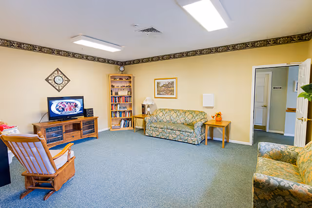 A cozy living room in an assisted living facility featuring two floral patterned sofas, a wooden rocking chair, a wooden TV stand with a television displaying a food image, a bookshelf filled with books, a side table with a lamp, and a framed picture on the wall. The room has beige walls, blue carpet, and fluorescent ceiling lights.