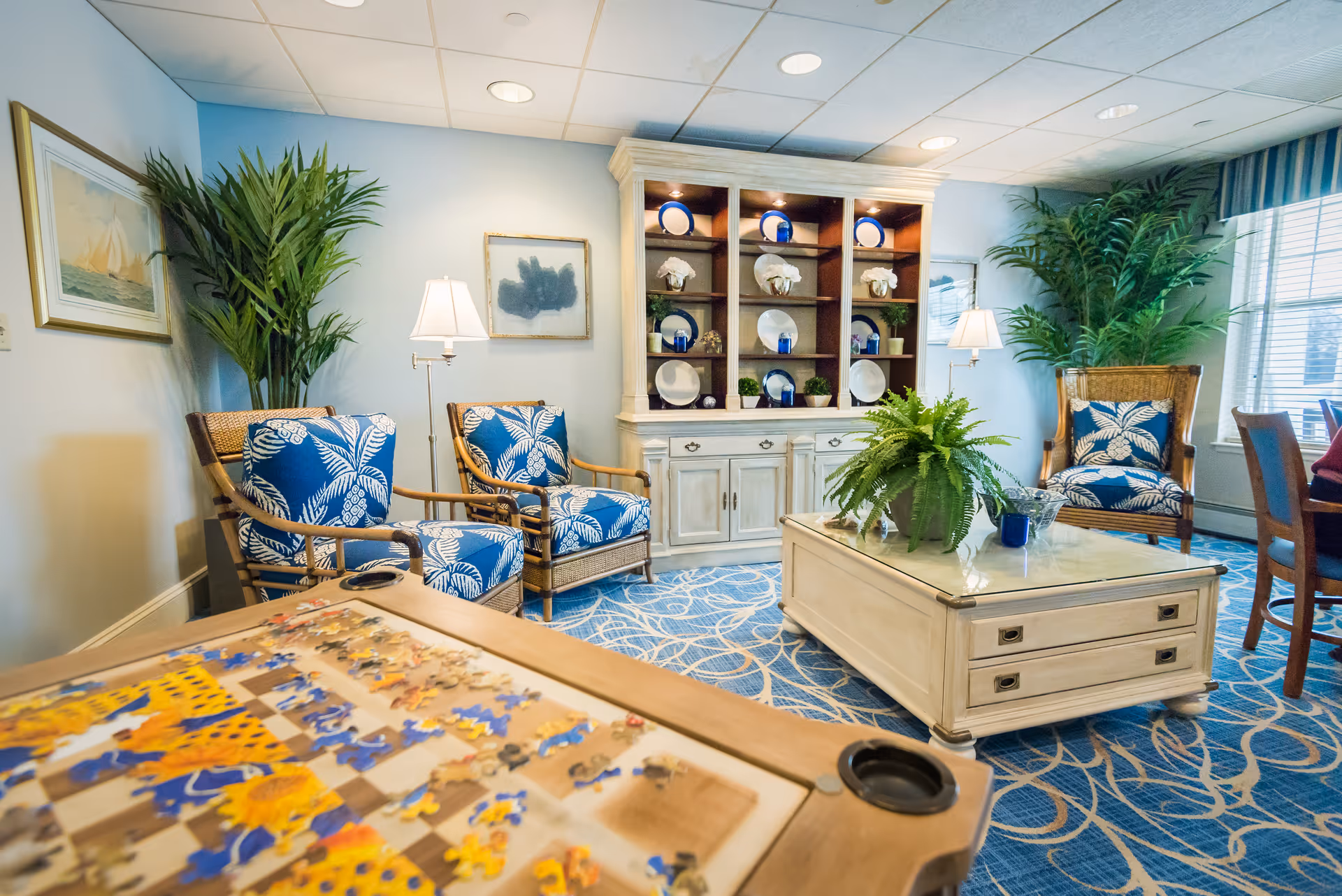 A cozy living room area with blue and white patterned armchairs, a white coffee table with a green potted plant, a wooden cabinet displaying decorative plates and figurines, and a puzzle table in the foreground. The room has blue carpet with a circular pattern, large windows with curtains, and several green plants adding a fresh touch.
