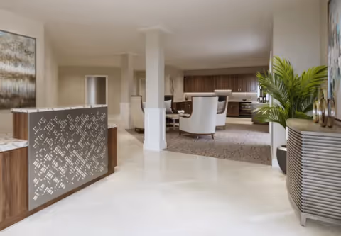 A spacious and modern interior area featuring a reception desk with a decorative front panel on the left, a seating area with white cushioned chairs and a round table in the middle, and a kitchen area with wooden cabinets and stainless steel appliances in the background. There is a large green plant on the right side next to a textured cabinet, and light-colored tiled flooring throughout the space.