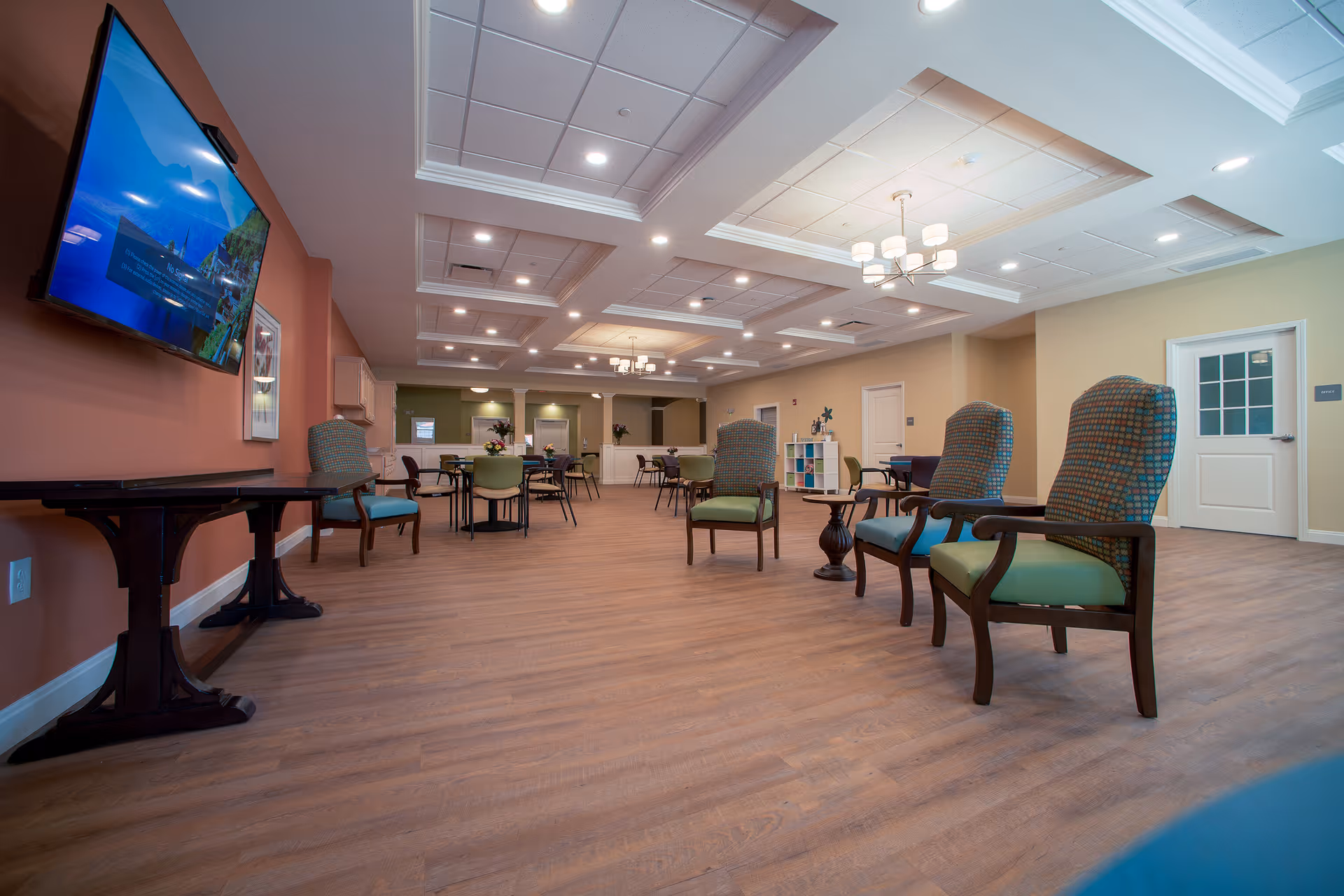 A spacious senior living common area with wooden flooring, multiple tables and chairs arranged for seating, a large flat-screen TV mounted on a peach-colored wall, and ceiling lights with decorative fixtures. The room has a warm and inviting atmosphere with beige and green walls and a white door on the right side.