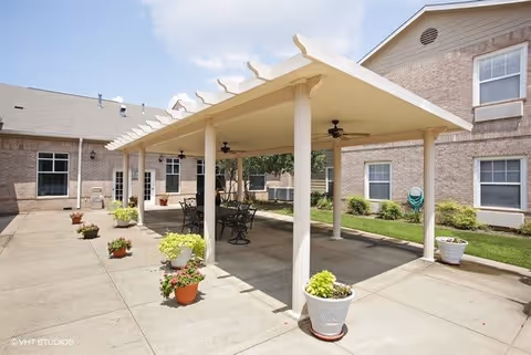 Outdoor patio area at Morada Pantego with a covered pergola supported by white columns, ceiling fans, and several metal tables and chairs. The patio is surrounded by a brick building with windows and doors, and there are potted plants placed around the concrete floor.