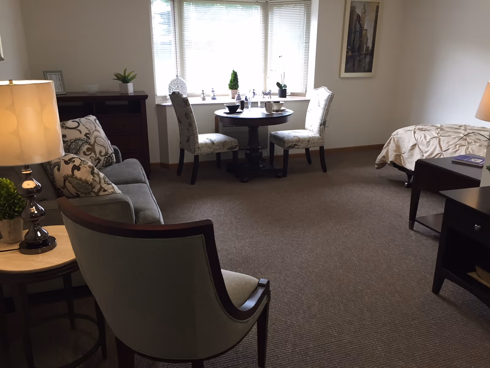 A cozy assisted living room featuring a small round dining table with two upholstered chairs near a window with blinds. There is a gray sofa with patterned cushions, a side table with a lamp and a small plant, and a bed with a beige comforter. The room has carpeted flooring and neutral-colored walls with a framed picture.