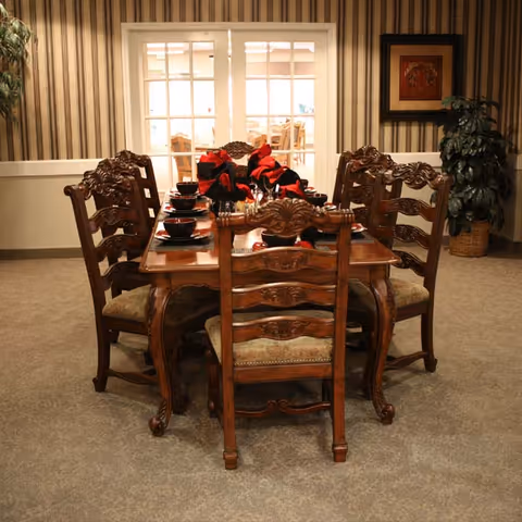 A dining room with an ornate wooden dining table and six matching chairs. The table is set with black bowls and plates, and decorated with red and black napkins. The room has striped wallpaper, a framed picture on the wall, a potted plant in the corner, and glass double doors leading to another room with more tables and chairs.
