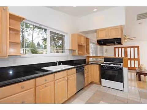 A bright kitchen with light wood cabinets, black countertops, a white sink under a large window, a stainless steel dishwasher, and a white stove with a black range hood. The kitchen opens into another room with a wooden door and a ceiling fan.